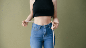 Pessoa medindo a cintura com fita métrica, vestindo top preto e calça jeans, em imagem que representa preocupação com a imagem corporal.